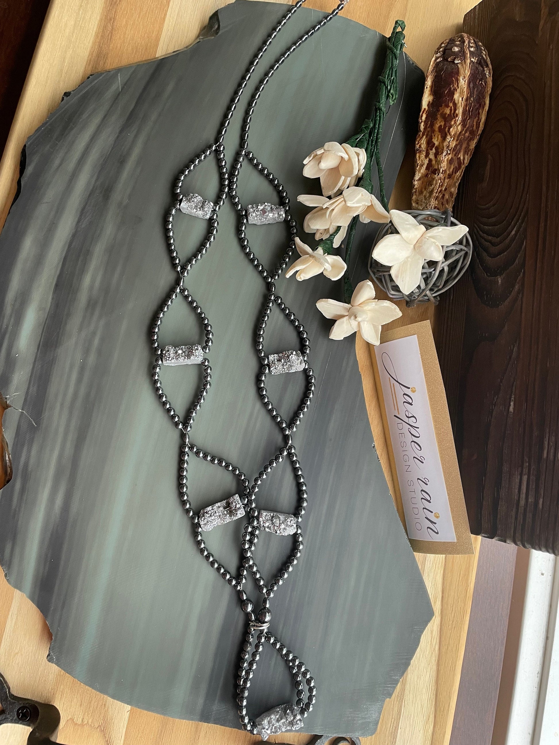 Platinum Druzy and Hematite long length necklace sitting on a stone tablet. Gold and white Jasper Rain brand logo sign is pictured.